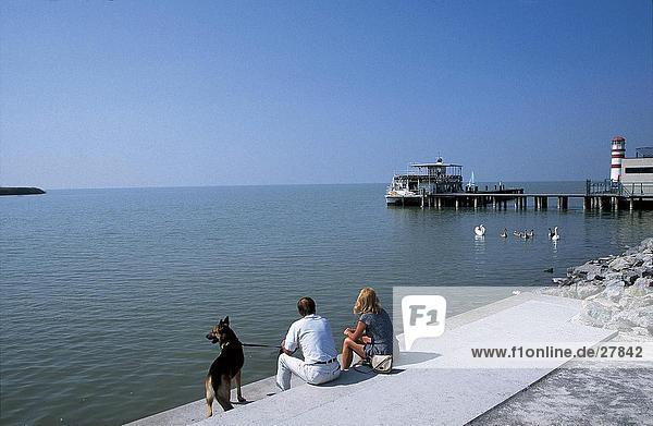 Paar mit Hund an der K??ste mit Pier im Hintergrund  Neusiedler See Lake  Podersdorf  Burgenland  Österreich