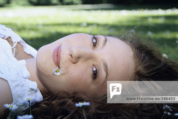 Junge Frau mit Gänseblümchen im Mund auf Gras liegend  Nahaufnahme  Portrait