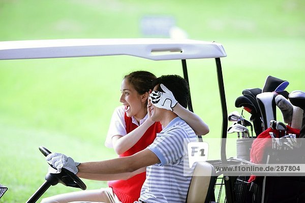 Eine Frau umfasst die Augen von ihrem Partner wie er ein Golf-Cart fährt