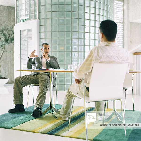 Zwei Männer sitzen im Gespräch von einer Tabelle in einer Büroumgebung.