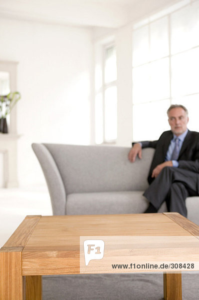 Erwachsener Mann sitzt auf dem Sofa und schaut weg (Fokus auf den Tisch im Vordergrund)
