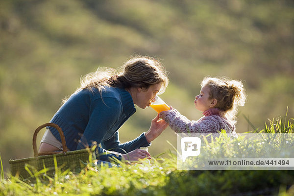 Mother sitting with daughter in meadow  drinking from daughters glass