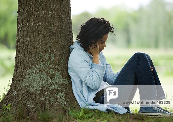 Junge Frau sitzt auf dem Boden und lehnt sich an den Baum.