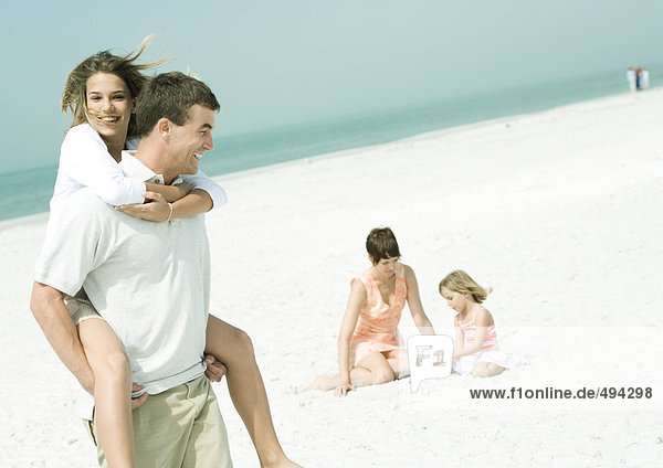 Familie am Strand  Mann mit Teenager-Tochter Huckepack  während Mutter mit Tochter auf Sand sitzt.
