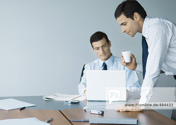 Businessman leaning on table with cup in hand while colleague uses laptop