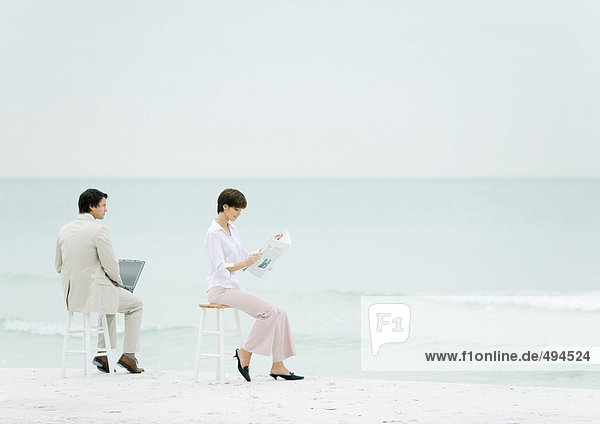 Businesspeople sitting on stools on beach  one using laptop  another reading newspaper
