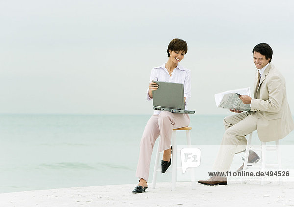 Geschäftsleute sitzen auf Hockern am Strand  einer mit Laptop  ein anderer mit Zeitung lesen