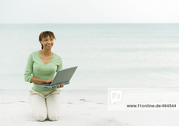 Frau am Strand  kniend auf Sand mit Laptop