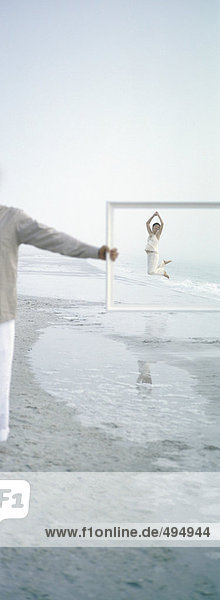 On beach  woman jumping in distance  seen through frame held by man