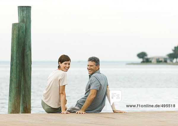 Couple sitting on edge of dock  smiling over shoulders at camera