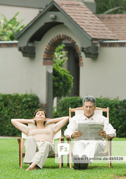 Couple sitting in deckchairs on lawn  man reading newspaper and woman napping