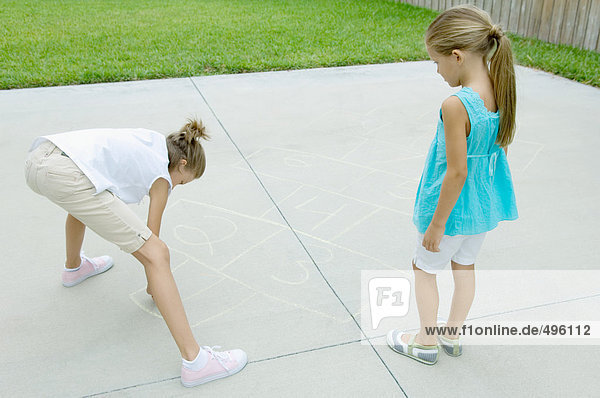 Girl drawing hopscotch game on driveway while second girl waits