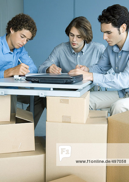 Three male colleagues working at table  cardboard boxes in foreground