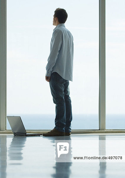 Mann stehend  Blick aus dem Erkerfenster  Laptop auf dem Boden neben ihm