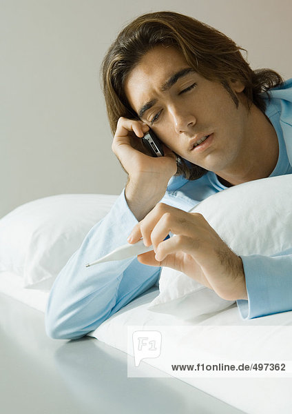 Mann liegt im Bett  benutzt Handy und hält Thermometer hoch