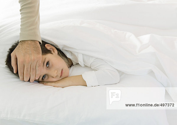 Kind im Bett liegend mit Vaters Hand auf der Stirn