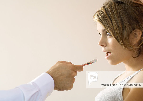 Arzt hält Zungenspatel zum Mund der jungen Frau hin.