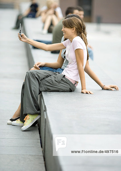 Zwei Teenager-Mädchen sitzen auf einer Bank in der Stadt und fotografieren mit dem Handy.