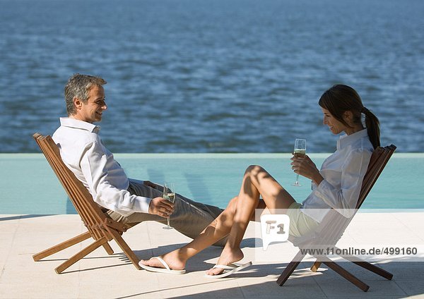 Paar sitzt in Liegestühlen am Meer und trinkt Champagner.