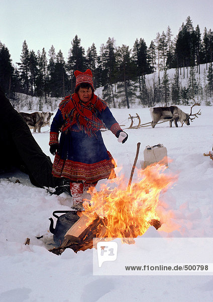 Finnland,  Saamifrau steht neben dem Feuer,  Schlittenrentier im Hintergrund
