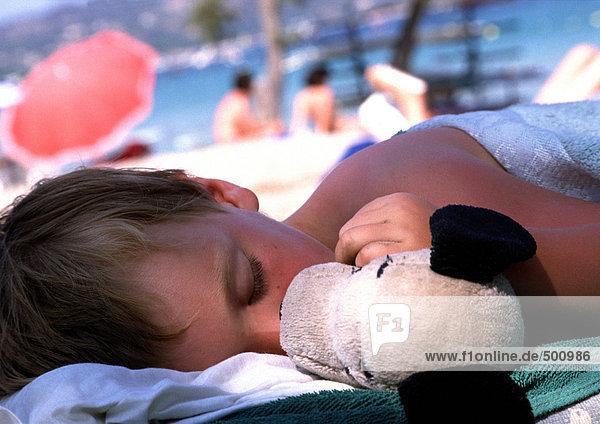 Kind schläft mit Puppe am Strand.