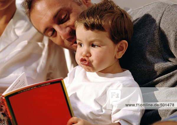 Kleines Kind macht ein albernes Gesicht  schaut sich ein Buch mit einem älteren Mann an.