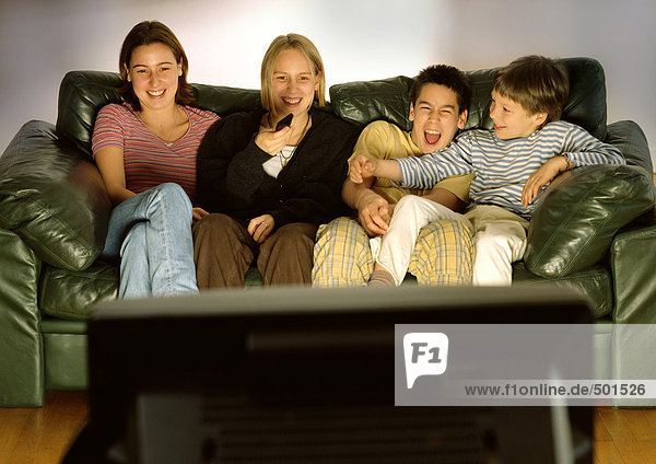 Drei Jugendliche und ein Kind sitzen gemeinsam auf der grünen Couch  lachend  verschwommene Rückansicht des Fernsehers im Vordergrund.