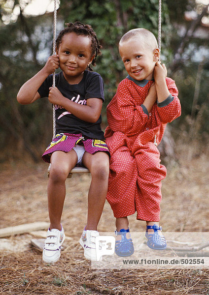 Kleiner Junge und Mädchen sitzen auf einer Schaukel  jede hält eine Seite des Schaukelseils  lächelnd vor der Kamera  volle Länge.
