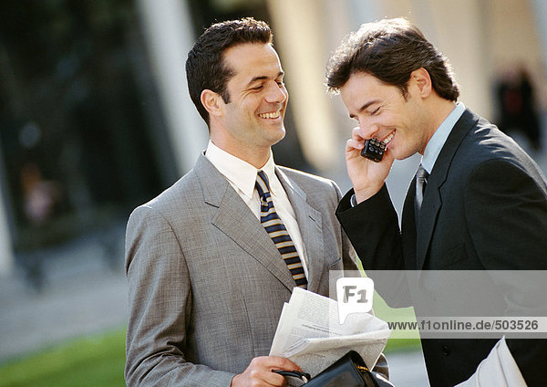 Two businessmen side by side outside  one using cell phone