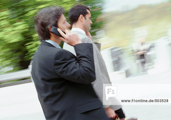 Two businessmen walking outside  one using cell phone  blurred