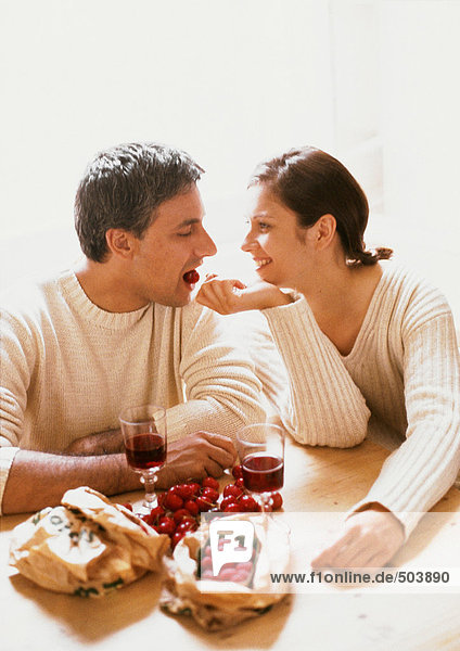 Paar am Tisch sitzend  Kopf und Schultern  mit Beeren  Kirschen und Gläsern Rotwein  Frau füttert Mann Kirsche