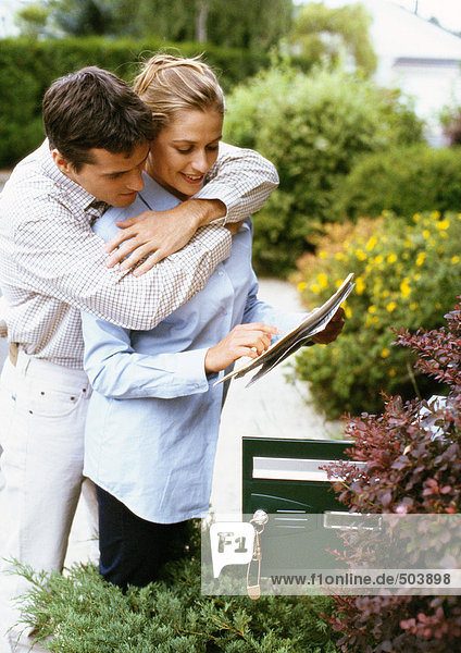 Frau hält Post  Mann umarmt sie von hinten  neben Briefkasten  Sträucher im Hintergrund