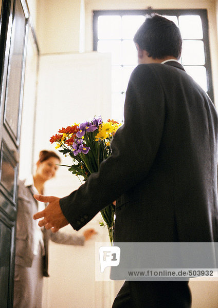 Mann mit Blumenstrauß  Frau lächelnd im Hintergrund  Rückansicht