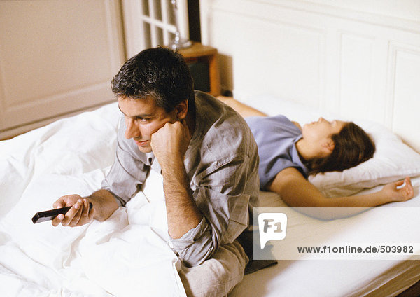 Mann sitzt im Bett,  zeigt auf die Fernbedienung,  Frau liegt hinter ihm.