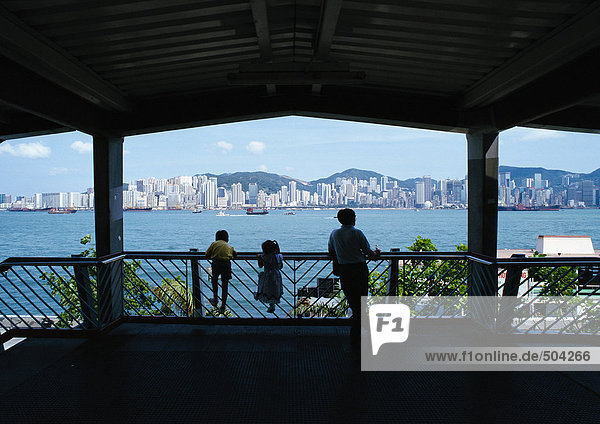 Hong Kong  silhouettes of adult with two children leaning on rail  rear view  city in distance
