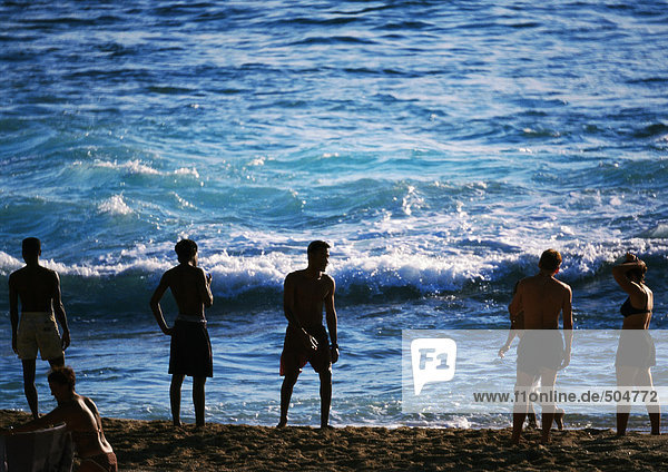 Silhouetten von Menschen  die am Strand am Rande des Meeres stehen  Wellen im Hintergrund
