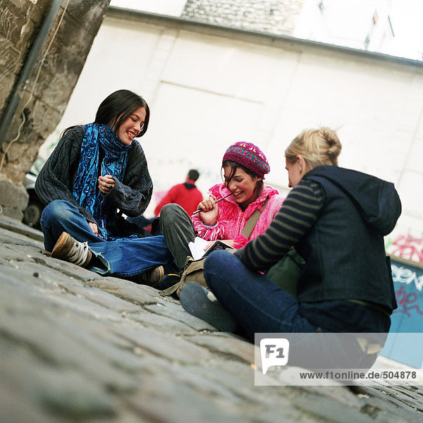 Drei Teenager-Mädchen sitzen auf Kopfsteinpflaster,  lächelnd