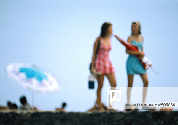 Zwei Frauen gehen am Strand  eine hält einen Sonnenschirm.