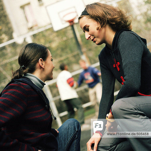 Junge Frauen sitzen zusammen  junge Männer im Hintergrund spielen Basketball.