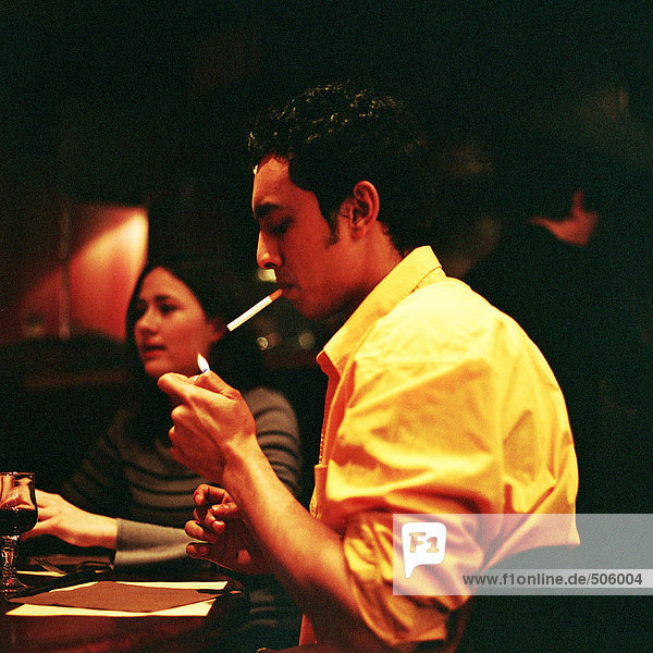 Junger Mann beim Anzünden einer Zigarette in einem Café  junge Frau im Hintergrund