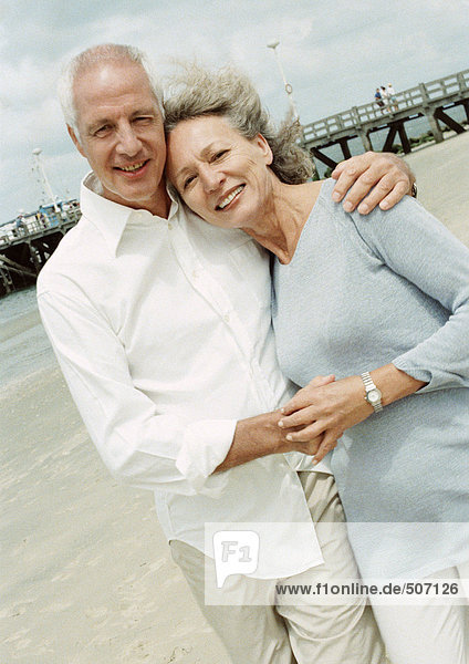 Ein reifes Paar mit Armen umeinander am Strand  lächelnd vor der Kamera.