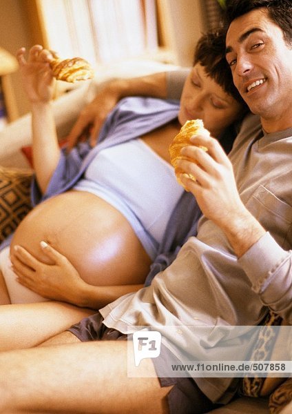 Man and pregnant woman eating croissants on sofa