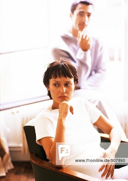 Schwangere Frau sitzt im Sessel,  Mann im Hintergrund
