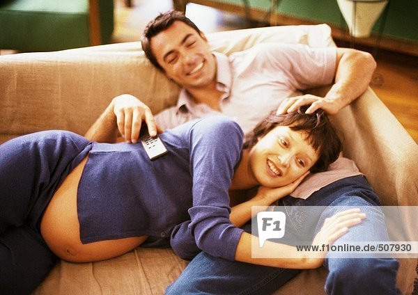 Man and pregnant woman on sofa  woman lying on man's lap  man holding remote control  portrait