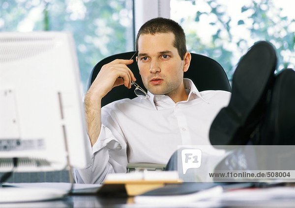 Geschäftsmann mit Brille neben dem Gesicht  Blick auf den Computerbildschirm  mit Füßen auf dem Schreibtisch sitzend