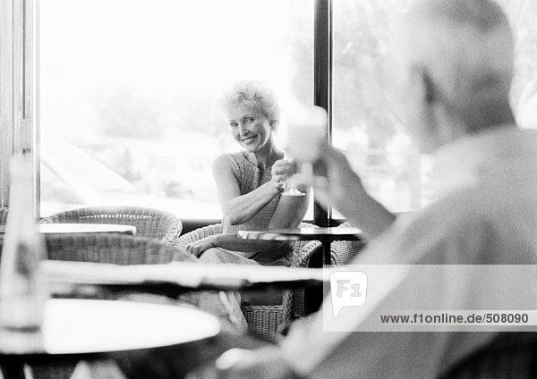 Mature man and woman looking at each other across the room in a cafe  raising glasses  blurred foreground  B&W