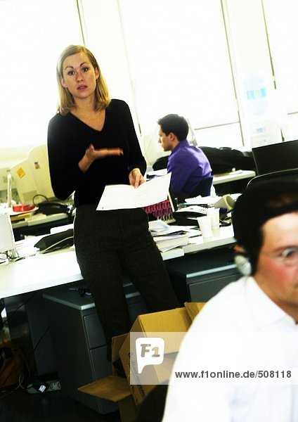 In office  woman standing with document  gesturing  other people working
