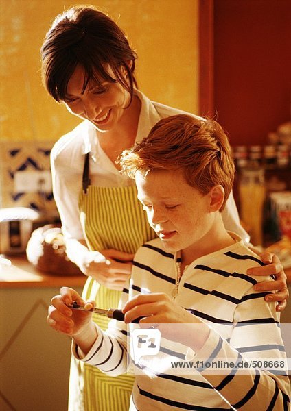 Frau lächelt in der Schürze mit den Händen auf der Schulter des Jungen  in der Küche