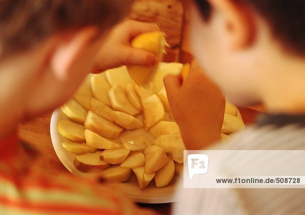 Zwei Kinder  eine quetschende Zitrone über geschnittenen Äpfeln  Kinder im Vordergrund verschwommen  Rückansicht