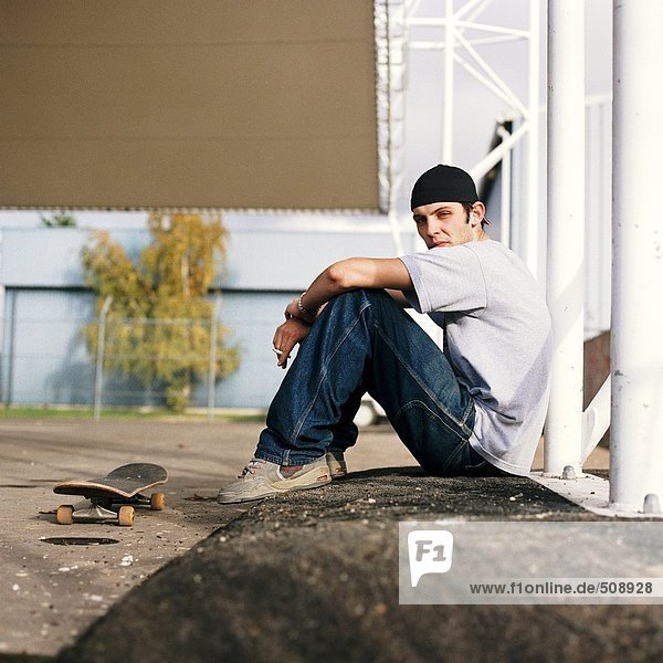 Junger Mann mit Skateboard im Freien sitzend  Seitenansicht  Portrait